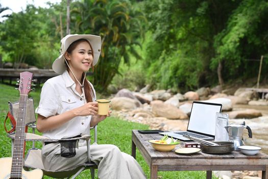 Happy female traveler drinking coffee, resting in folding chair near the river bank. Adventure, travel and camping concept.