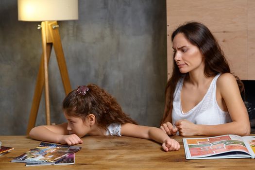 Portrait of a mother helping her small sweet and cute daughter to make her homework indoors. Mom is looking angry and her daughter is turned away.