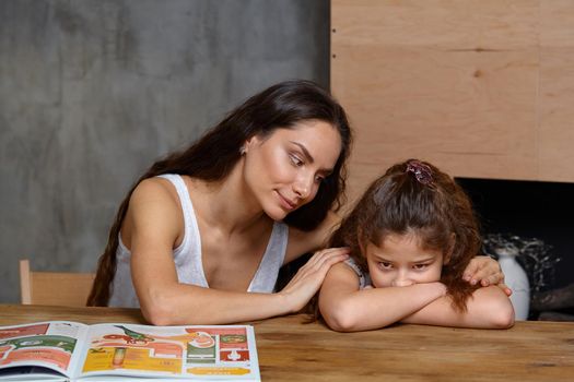 Portrait of a mother helping her small sweet and cute daughter to make her homework indoors. Mom is comforting her daughter.