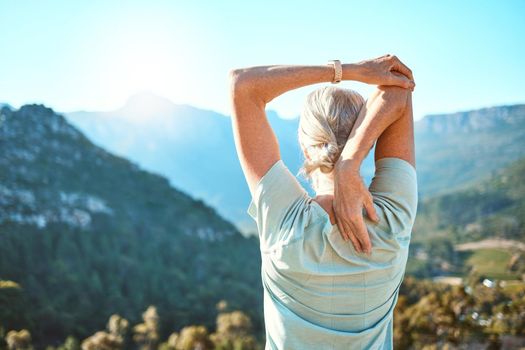 Rear view of a senior woman with grey hair stretching with her hands behind her head while standing outdoors and overlooking a scenic mountain view. Living healthy active lifestyle.