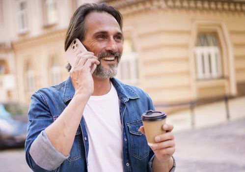 Happy middle aged man with grey bearded talking on the phone holding coffee in disposable paper cup standing outdoors in old city background wearing jeans shirt. Freelancer traveling man.