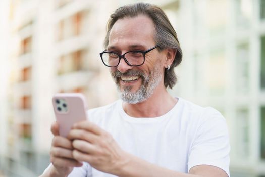 Happy caucasian middle aged man with grey beard reading or texting using smartphone standing outdoors city streets background wearing white shirt. Middle aged man on street of european city.