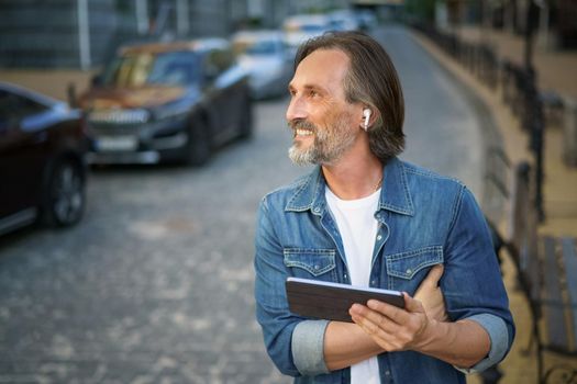 Middle aged freelancer man use gps or calling digital tablet while walking on urban streets. Mature man use wireless earphones talking or listening music while travel in old town streets.