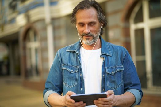 Playing games while traveling handsome mature man use digital tablet standing outdoors urban city streets. Mature man listening music use wireless earphones while travel old town streets.