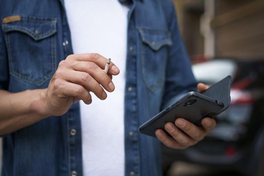 Close up. Unhealthy addiction smoking cigaret man read social media using his smartphone wearing jeans shirt and white t-shirt standing outdoors on urban city background. No face visible.