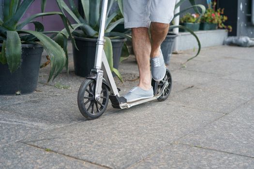 Close up male feet in sneakers shoes stand on scooter with big wheels having a ride on the streets or park after work outdoors with agave plants on background. No face visible. Focus on front wheel.