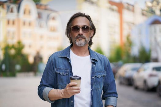 Handsome middle aged man with grey beard walking in the city holding take away coffee in paper cup. Handsome man traveling enjoying free time. Creative man on a coffee break outdoors.