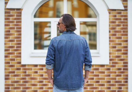 Back view of a man standing alone looking sideways at old town building while traveling in european cities during vacation time wearing denim jeans shirt. Travel concept.