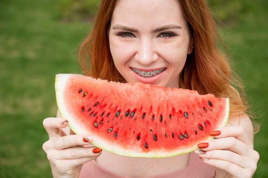 Beautiful red-haired woman smiling with braces and about to eat a slice of watermelon outdoors in summer.