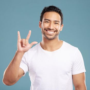 Handsome young mixed race man gesturing rock on while standing in studio isolated against a blue background. Hispanic male showing the sign language of I love you to show affection or romance.