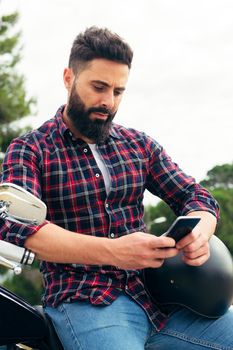 vertical photo of a handsome bearded man consulting his mobile phone sitting on his motorcycle with his helmet under his arm, concept of freedom and biker lifestyle, copy space for text