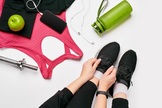 Young woman in sporting leggings laces sneakers, preparing for training. Accessories for sports, bottle of water, smartphone, headphones, fitness bracelet on white background flat lay top view. Fitness concept