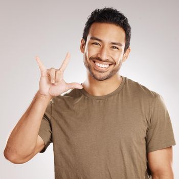Handsome young mixed race man gesturing rock on while standing in studio isolated against a grey background. Hispanic male showing the sign language of I love you to show affection or romance.