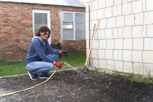 Caucasian woman wearing gloves crouching while watering seeds on the soil using a hose