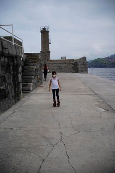 girl skating with inline skates with a woman walking in the background of the marina of castrourdiales in cantabria