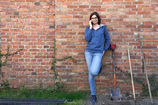 Portrait of a woman talking to the mobile while leaning in a brick wall next to rural work tools