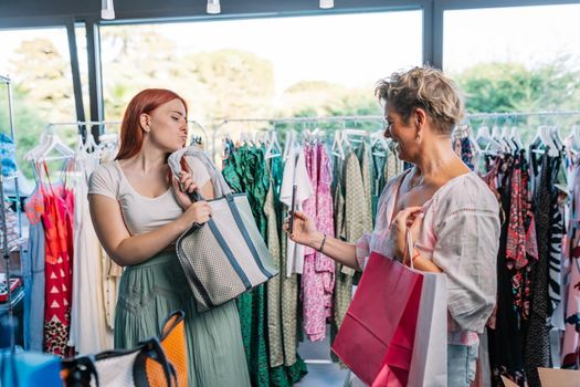 two women friends shopping in a clothing shop, taking pictures with their smartphones for social media. mother and daughter enjoying a day of shopping. shopping concept. natural light from window.