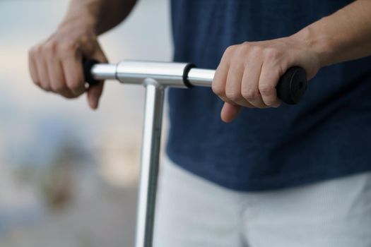 Close up man hands holding scooter handle ride on the streets or park after work outdoors wearing dark blue t-shirt and light shorts. No face visible. Selective focus on left hand.