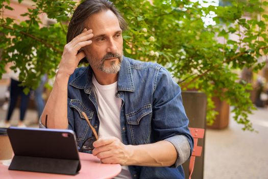 Handsome middle age freelancer man working outdoors using digital tablet planing project. Middle aged man looking away sitting at cafe or street restaurant during vacation traveling.