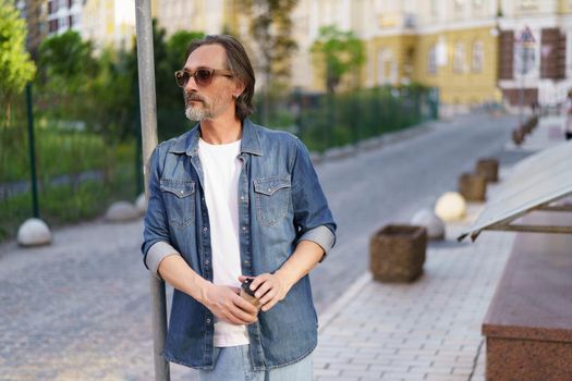 Emotionally exhausted middle age crisis mature man leaned on road sign holding paper cup off coffee. Experiencing hangover middle age alcoholic man drink using paper cup to hide alcohol.