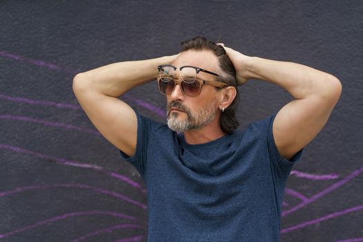 Middle aged man put hands behind head with glasses and sunglasses on his face standing next to dark urban wall wearing dark blue t-shirt. Mature business man or freelancer outdoor.