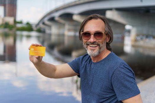 Middle aged man showing debit, credit card sitting outdoors next to river working using digital tablet pc, shopping online or paying online banking. Online banking concept. Finance concept.