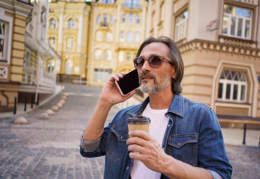 Talking on the phone middle aged silver beard man holding cup of coffee using in disposable paper cup standing outdoors in old city background wearing jeans shirt. Freelancer traveling man.