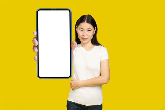 Showing phone white screen middle aged asian woman 40s holding smartphone mobile app advertisement and excited smile on camera isolated on yellow background. Product placement.