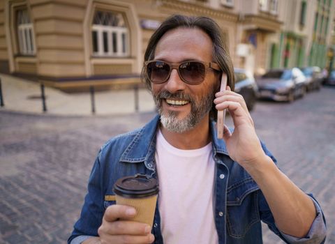 Close up portrait talking on the phone middle aged bearded man with take away coffee in paper cup standing outdoors in old town wearing jeans shirt. Freelancer traveling man. Business on the go.