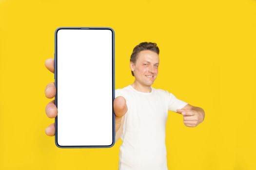 Huge smartphone in hand of handsome blond man pointing at white empty screen, wearing white t-shirt and jeans isolated on yellow background. Mobile app advertisement, great offer.