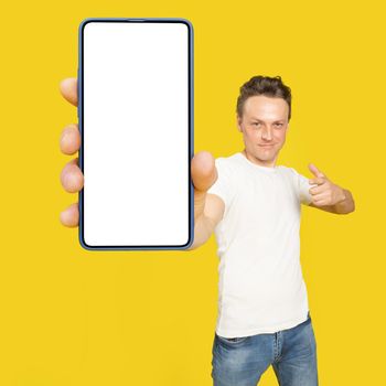 Handsome man holding huge smartphone with white screen pointing at white empty screen, wearing white t-shirt and jeans isolated on yellow background. Mobile app advertisement, great offer.