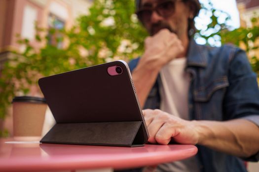 Low view middle aged man working outdoors using digital tablet drinking coffee. Handsome freelancer traveling man checking work or social media sitting at city street cafe. Selective focus on tablet.