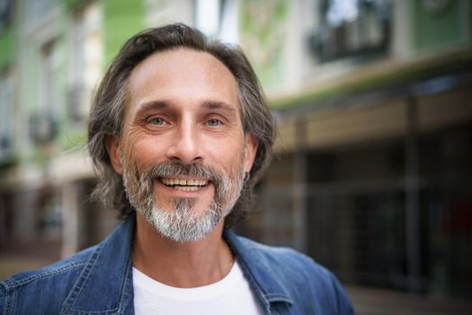 Happy handsome middle aged man standing outdoors on street of city looking at camera with a charming smile. Middle aged grey bearded man traveling in city smiling on camera wearing casual.