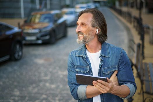 Middle aged freelancer man use gps or calling digital tablet while walking on urban streets. Mature man use wireless earphones talking or listening music while travel in old town streets.