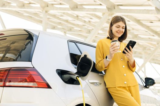 Vehicle charging at public charging station near solar power plant. Woman with a mobile phone and coffee cup standing near charging electric car. Eco friendly alternative energy transport.