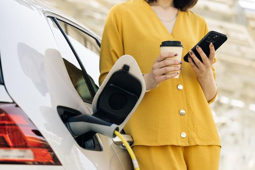 Unrecognizable caucasian woman using smart phone and waiting power supply connect to electric vehicles for charging the battery in car. Plug charging an Electric car from charging station.