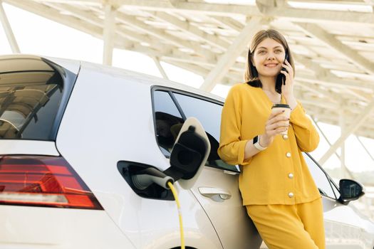 Attractive business woman taking phone call standing near her electric car. Positive woman have talking conversation by phone near her electric car and waits when the vehicle will be charged.