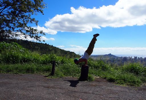 Man wearing beanie, muscle t-shirt, yoga pants, and slippers does balances on hands as he does Mayurasana or Peacock Pose on tree stump on Tantalus mountain of Oahu with Honolulu cityscape in the distance on a beautiful day.