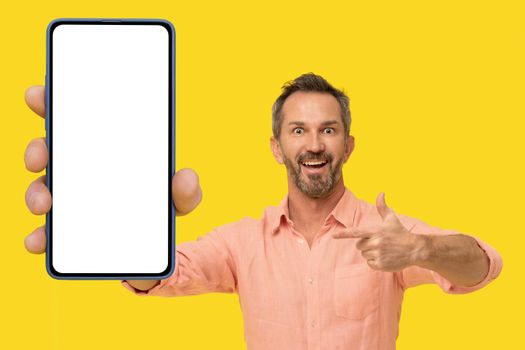 Middle aged handsome grey haired man pointing at smartphone in hand amazed smiling looking at camera wearing peach shirt isolated on yellow. Mature fit man with phone app advertisement.