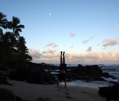 Man Handstanding on beach at dawn as wave crash on the North Shore and moon in the sky of Oahu, Hawaii.                    