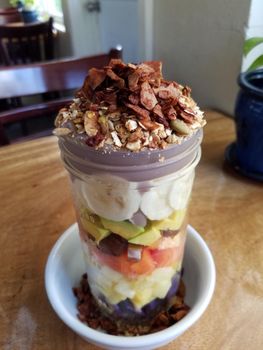 Poi-fect Parfait featuring Layers of fresh fruit (papaya, banana, pineapple, ʻūala, and avocado when available) topped with local poi and our house-made granola, toasted cacao coconut flakes and honey.  In a glass cup on top a small white plate on a wood table. Itʻs like eating a rainbow!
