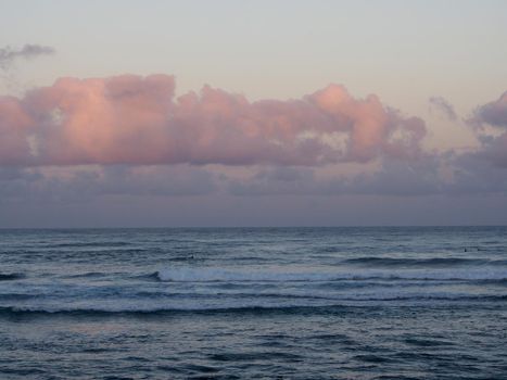 Beautiful pink and purple Dawn over the ocean with waves rolling towards the shore on the North Shore of Oahu with surfers in the water.    
