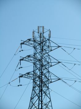 High Voltage Power Lines intersect at a large metal Utility pole with cell tower against a blue sky.