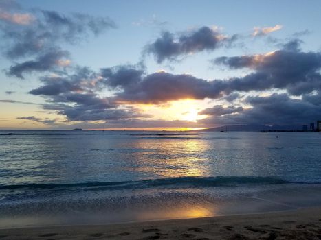 Dramatic Sunset over the clouds and reflecting on the Pacific ocean with boats on the water on Waikiki beach on Oahu, Hawaii with Waianae Mountain range visible.