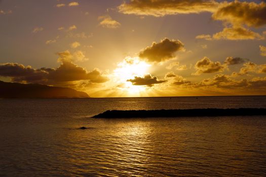 Beautiful Sunset over the ocean through the clouds with jetty in the water at Hale'iwa Beach Park on the North Shore of Oahu.    