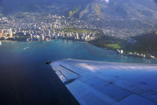 Aerial high in the sky shot of window view of plane leaving Honolulu, Hawaii with the wing of a commercial jet plane with large ship in the water and Diamondhead, Waikiki, beach, and Kapiolani park of Oahu visible.