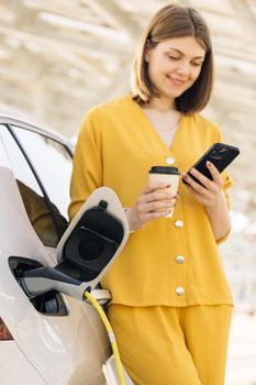 Caucasian woman with a mobile phone and coffee cup standing near charging electric car. Vehicle charging at public charging station near solar power plant. Eco friendly alternative energy transport.