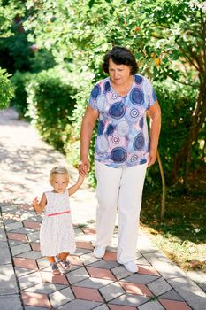 Grandmother leads little girl by the hand along the paving stones in the park. High quality photo