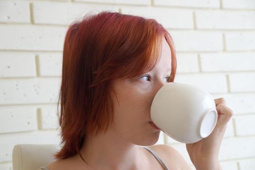 red-haired teenage girl drinks from a white cup of coffee and looks out the window.