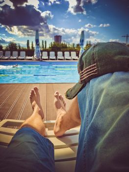 Men's legs in denim shorts on a sun lounger in front of the pool on a sunny day. Stylish cap on the knee